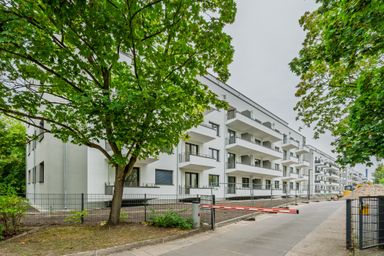 Modern white apartment building with balconies, surrounded by green trees and a fenced path under construction on a cloudy day.
