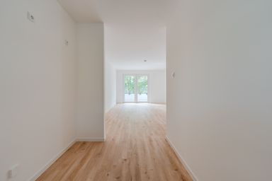 A bright, empty room with light wood flooring, white walls, and a large window facing a leafy view.