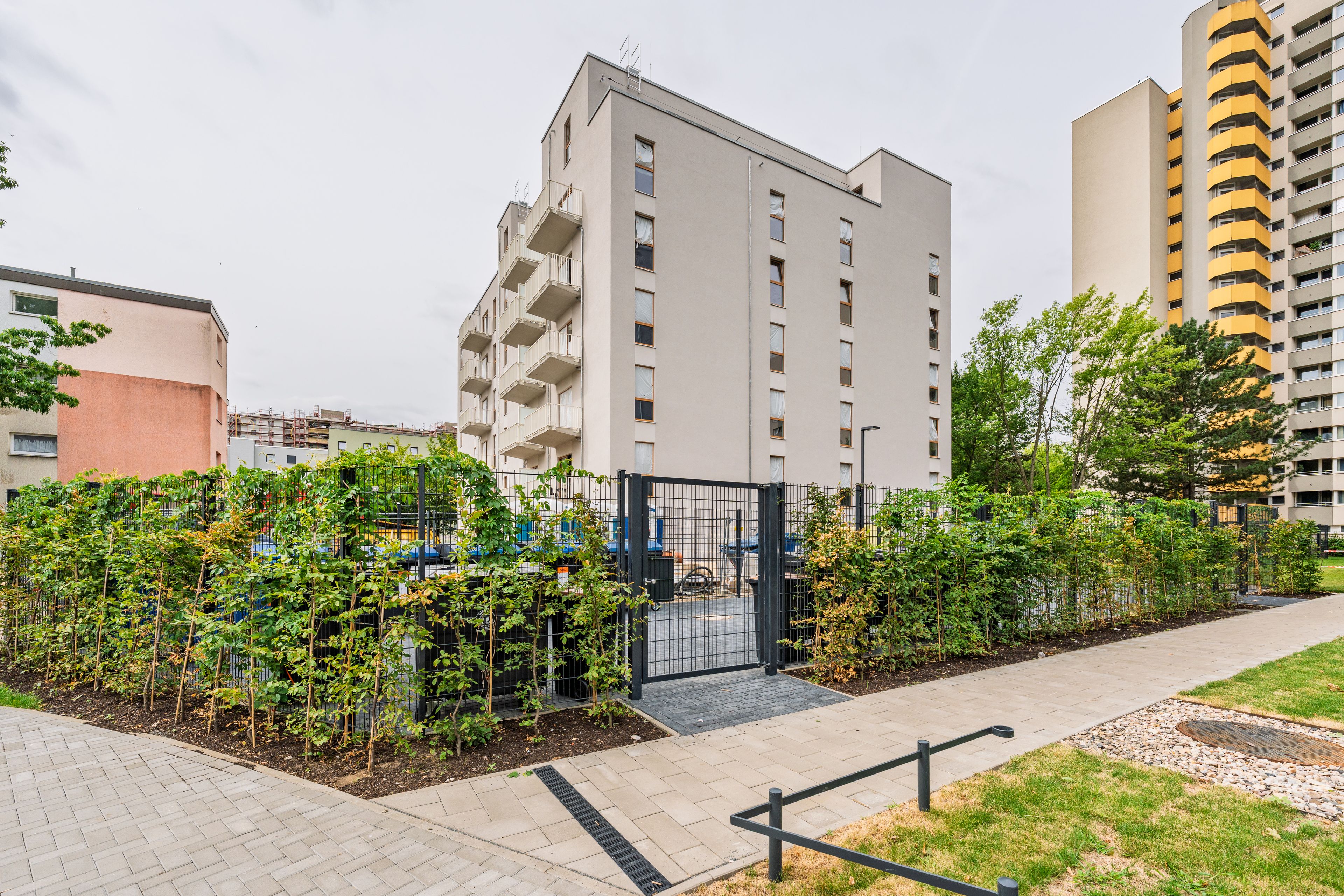 A modern apartment building with balconies, surrounded by greenery and a gated fence, in an urban residential area with additional high-rise buildings nearby.