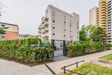 A modern apartment building with balconies, surrounded by greenery and a gated fence, in an urban residential area with additional high-rise buildings nearby.