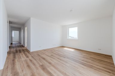 Leerer, heller Raum mit Holzoptikboden, weiß gestrichenen Wänden und einem Fenster, das Tageslicht hereinlässt.