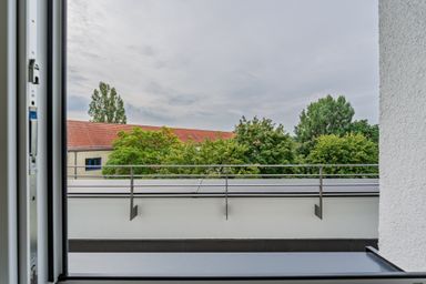 View from a balcony through an open window, showing a red-roofed building and green trees under a cloudy sky.