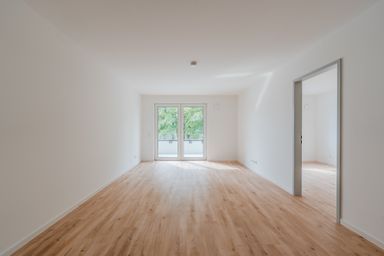 Empty room with light wooden floors, white walls, and a large window with a view of green trees. A doorway leads to another room.