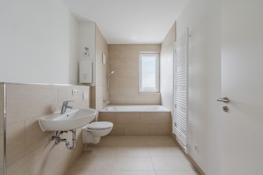 Modernes Badezimmer mit Badewanne, Waschbecken, Toilette und Handtuchheizkörper, beige Fliesen und Fenster.