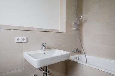 Modern bathroom with a white sink, chrome faucet, bathtub, and showerhead against beige tiled walls. Electrical outlets are visible.