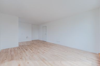 Empty room with light wood flooring, white walls, and a closed white door. The room is spacious and well-lit.