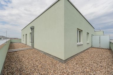 Flachdach eines elfstöckigen Gebäudes mit Kiesbelag und grüner Fassade, zwei Fenstern und einer Tür.