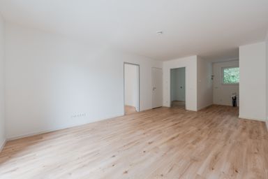 Empty room with light wood flooring, white walls, and multiple doorways. A window on the right provides natural light.