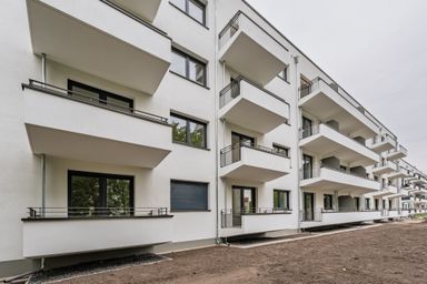 Modern apartment building with multiple balconies, white facade, and large windows, overlooking a barren ground area.
