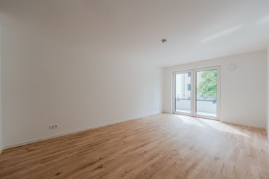 Empty room with light wood flooring, white walls, and a large glass door opening to a balcony, letting in natural light from outside.