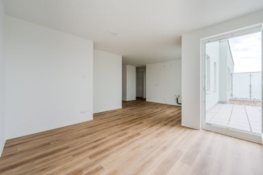 Leerer Wohnraum mit hellem Holzfußboden, weißen Wänden und Zugang zu einem Balkon mit Glasgeländer.