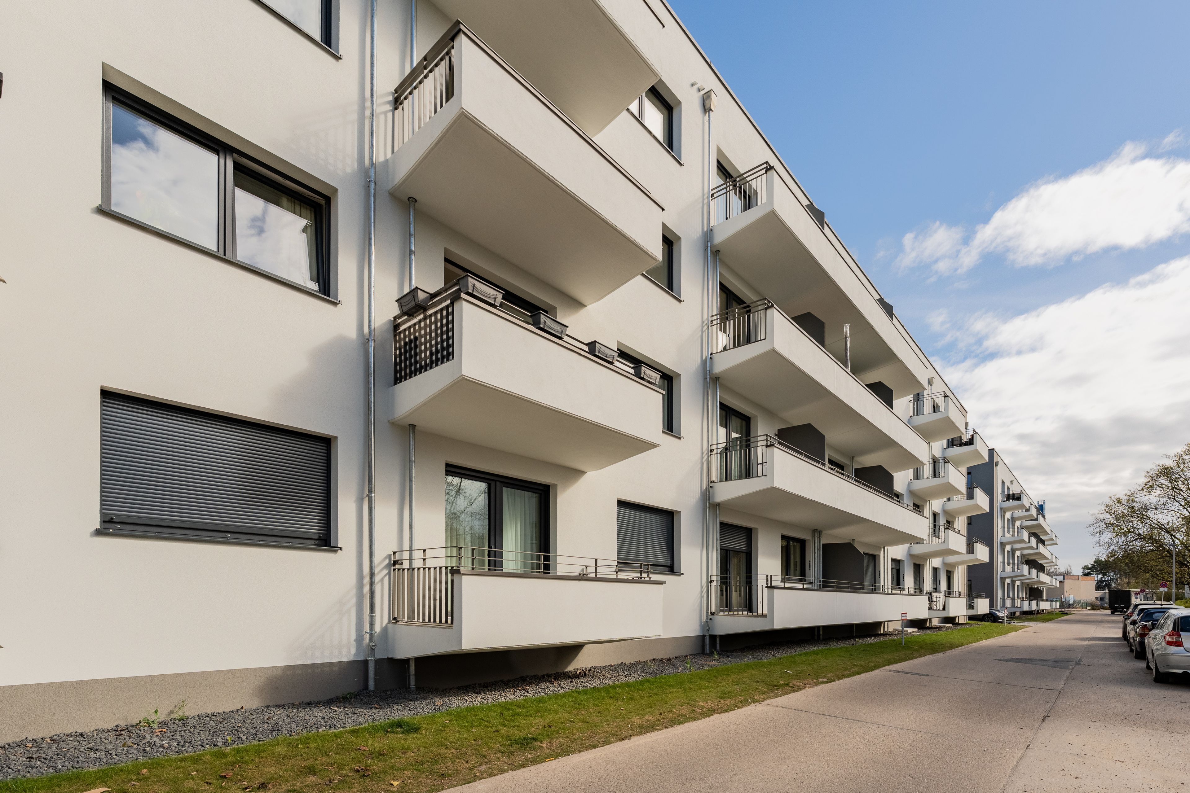 Modern white apartment building with multiple balconies, large windows, and a clear blue sky. Cars parked along the street.