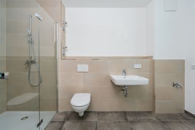 Modern bathroom with beige tiles, featuring a glass shower, wall-mounted toilet, and a white sink with a chrome faucet.