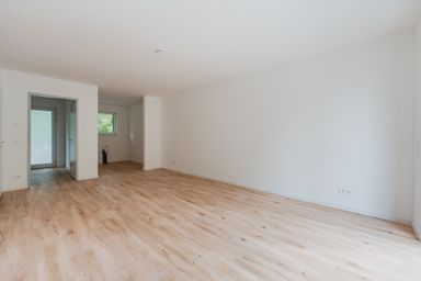 Empty room with white walls, light wood flooring, a doorway on the left, and a small window. Bright and spacious interior.