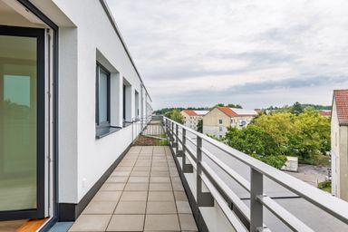 A long balcony with railings on a modern building overlooks a view of houses with red-tiled roofs and green trees under a cloudy sky.