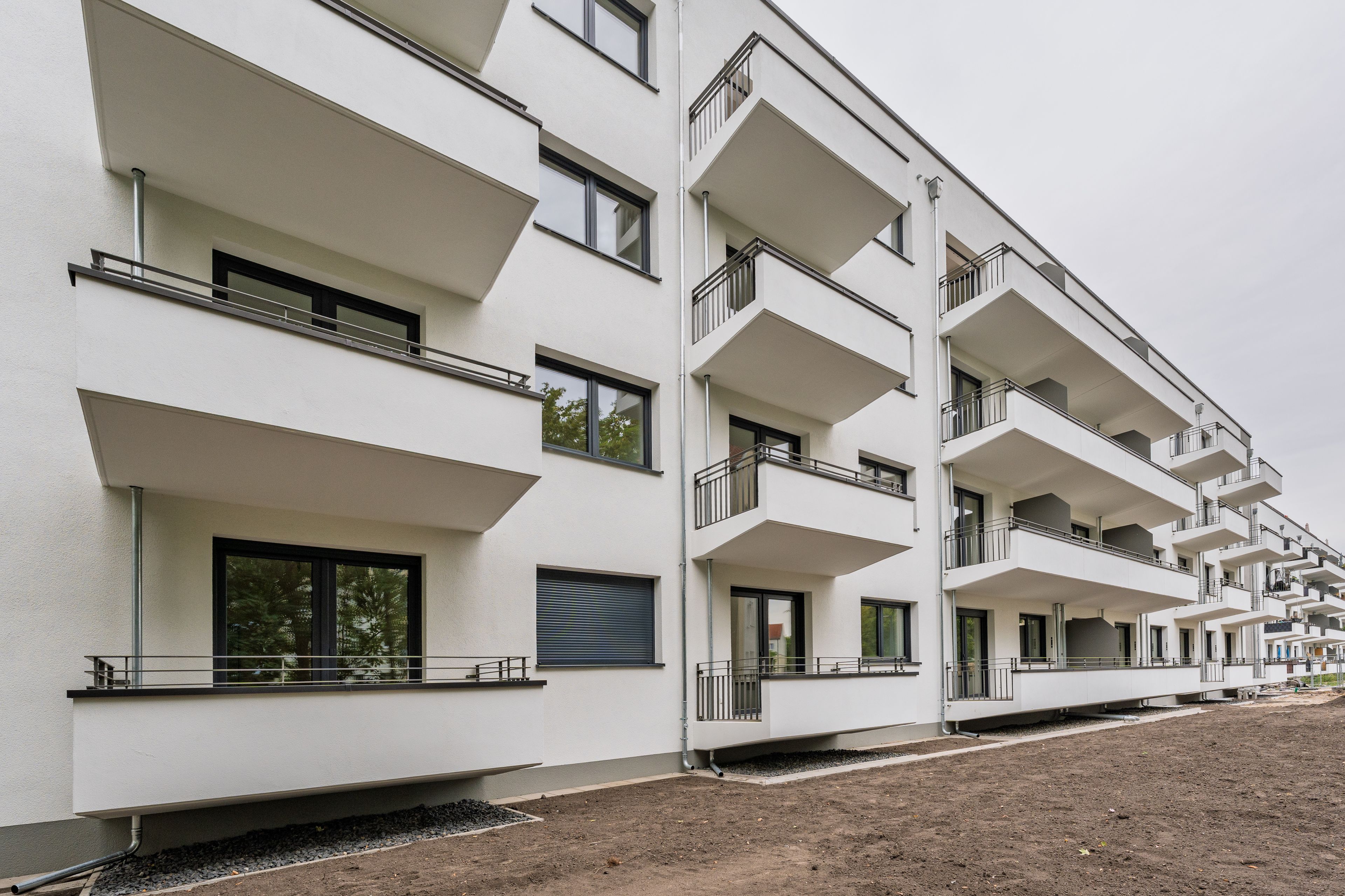 A modern apartment building with white facade, multiple balconies, and large windows against an overcast sky.