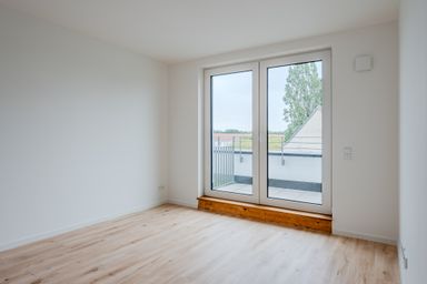 Empty room with light wooden floor, white walls, and a large glass door leading to a small balcony with a view of trees and rooftops.