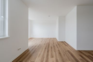 Leerer Raum mit Holzoptik-Boden und weißen Wänden, Blick auf Fenster links.