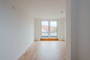 Empty room with light wood flooring, white walls, and a large window overlooking rooftops and a tree.