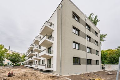 Newly constructed apartment building with multiple balconies and a gray and white facade, surrounded by trees and construction site.