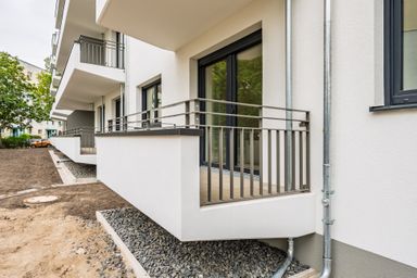 Modern apartment building with multiple balconies and sliding glass doors, surrounded by bare dirt and some greenery.