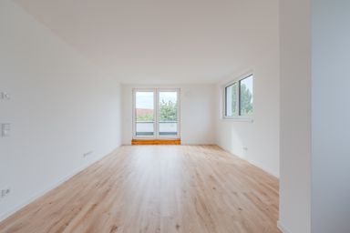 Empty room with light wood flooring, white walls, large windows, and a glass door leading to a small balcony with a view of trees and rooftops.