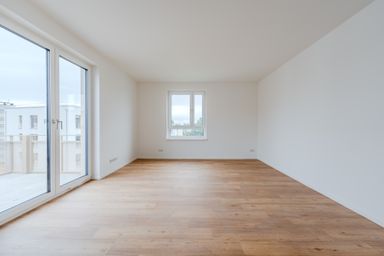 Leerer Raum mit Holzfußboden, großen Fenstern und Zugang zu einem Balkon, in einem Neubau.