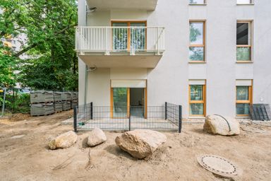 A modern apartment building exterior with a balcony and fenced patio on a dirt yard with large stones and stacked building materials nearby.