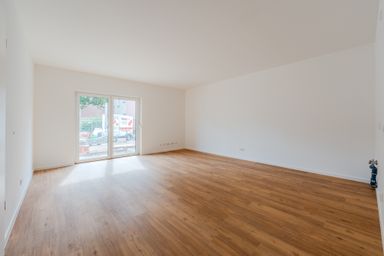 Empty room with white walls, large sliding glass door, and light wood flooring. Natural light enters from the window, revealing an outdoor view.