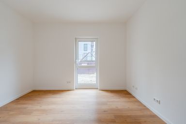 Empty room with white walls and wooden floor, featuring a single window with a view of a building.