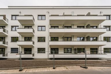 Modern apartment building with multiple balconies, large windows, and a fenced-off dirt area in front, set against a cloudy sky.