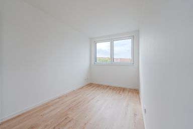 Empty room with white walls and light wood flooring, featuring a double window with a view of rooftops and trees.