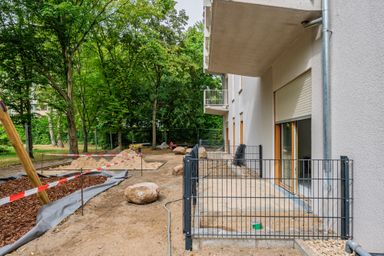 Construction site near a building with fenced walkways, sand, and wood chips, surrounded by trees. Equipment and barriers are visible.