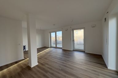 Leerer Raum mit Holzboden, weißen Wänden und großen Fenstern, die auf eine Terrasse führen.