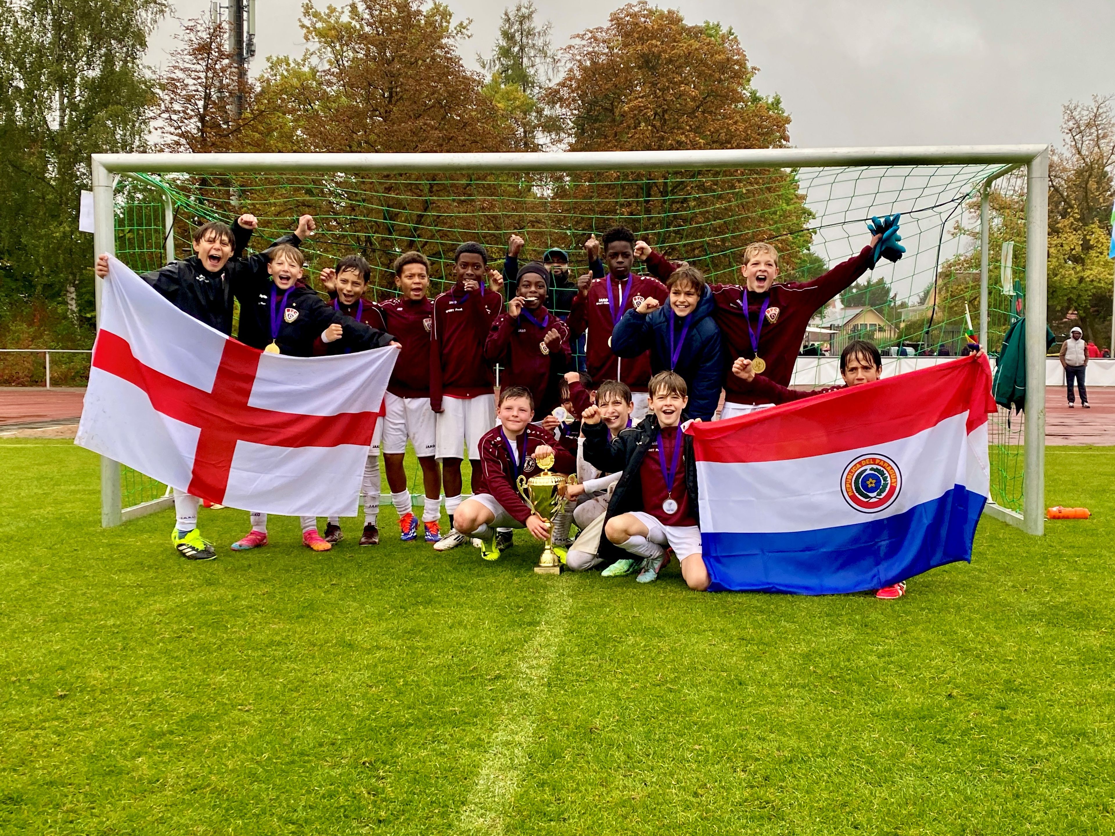 A group of young soccer players celebrating with medals and a trophy, holding English and Paraguayan flags, in front of a soccer goal.