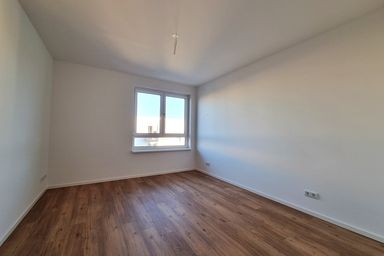 Leerer Raum mit Holzfußboden, weiß gestrichenen Wänden und einem Fenster, das Tageslicht hereinlässt.