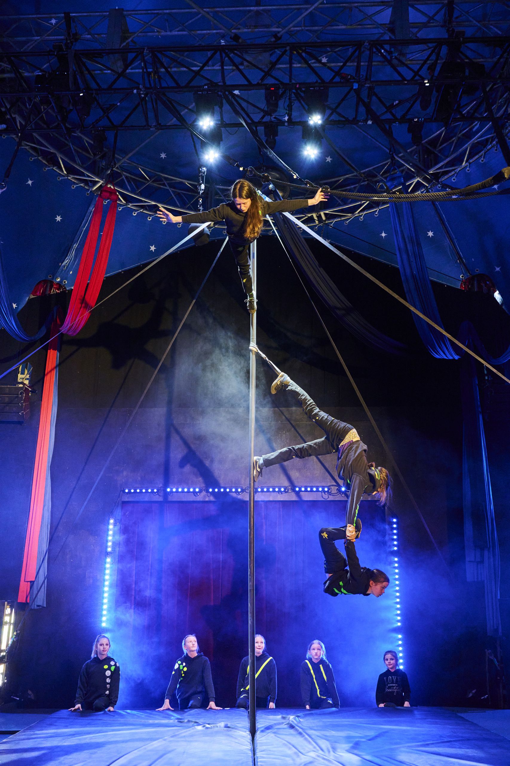 Zwei Akrobaten führen Kunststücke an einer Stange vor, während Zuschauer im Hintergrund sitzen.