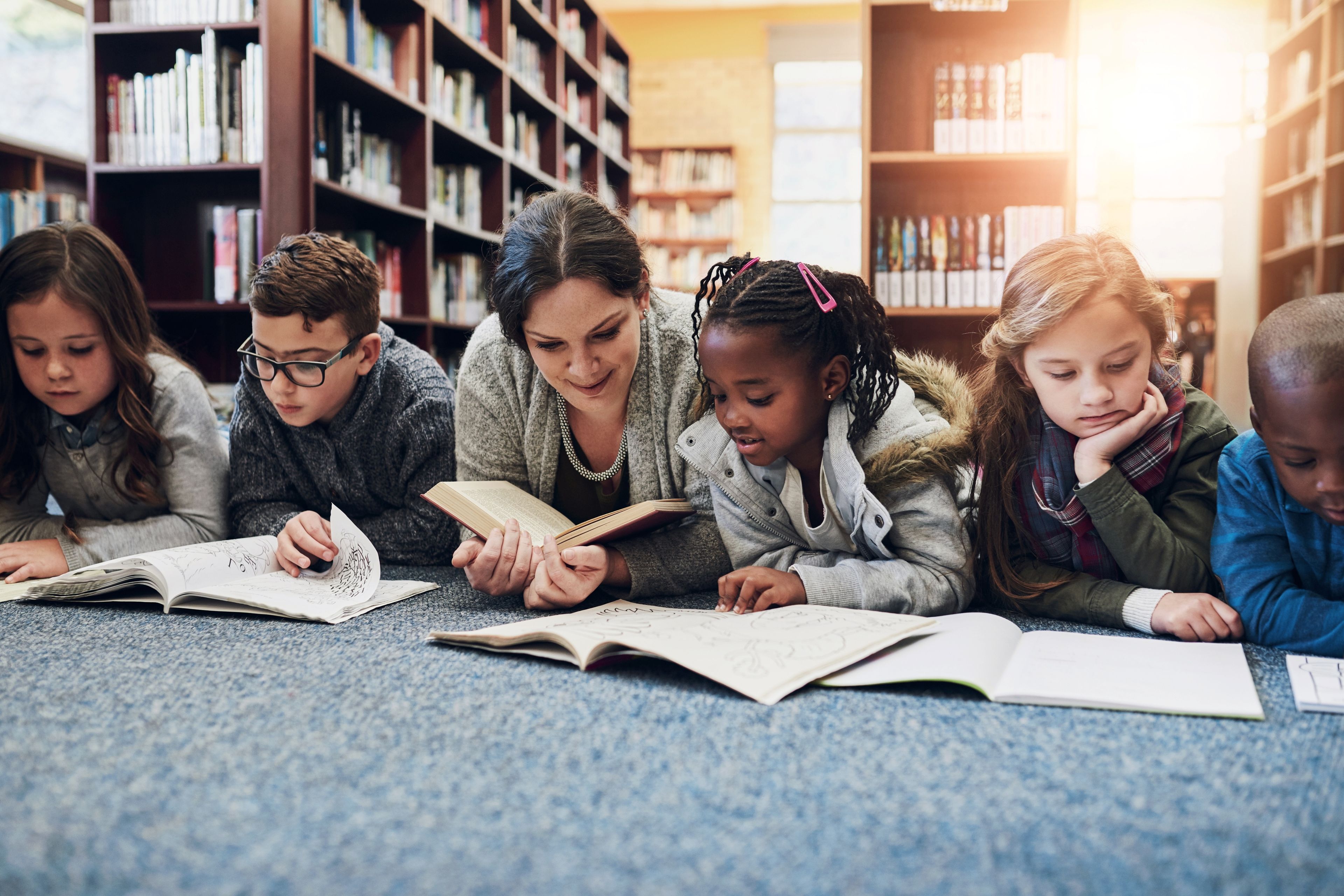 Eine Gruppe von Kindern und eine Frau liegen auf dem Boden einer Bibliothek und lesen Bücher.