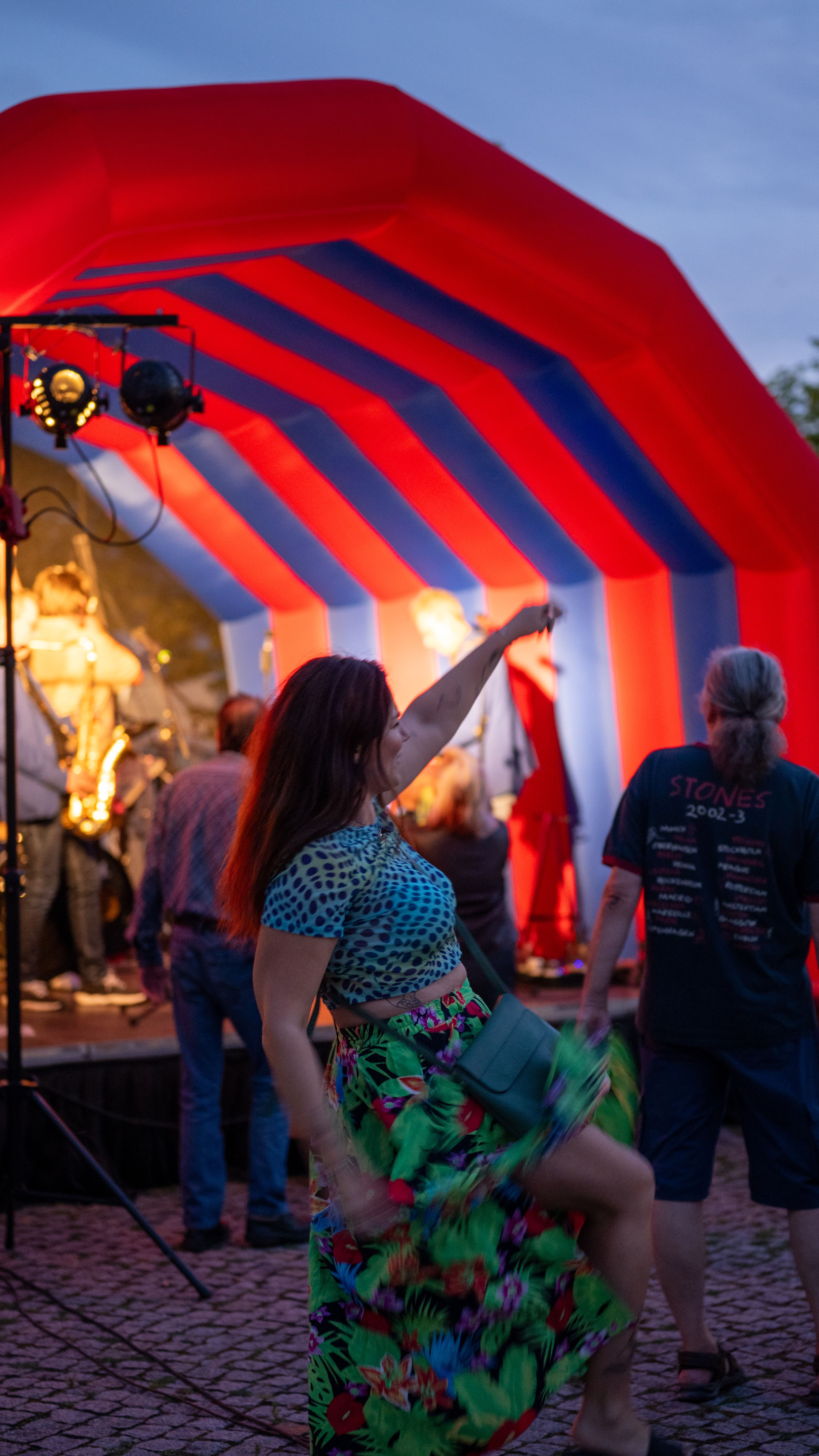 A lively outdoor concert with a woman dancing energetically near a stage with musicians under a red-and-blue striped tent.