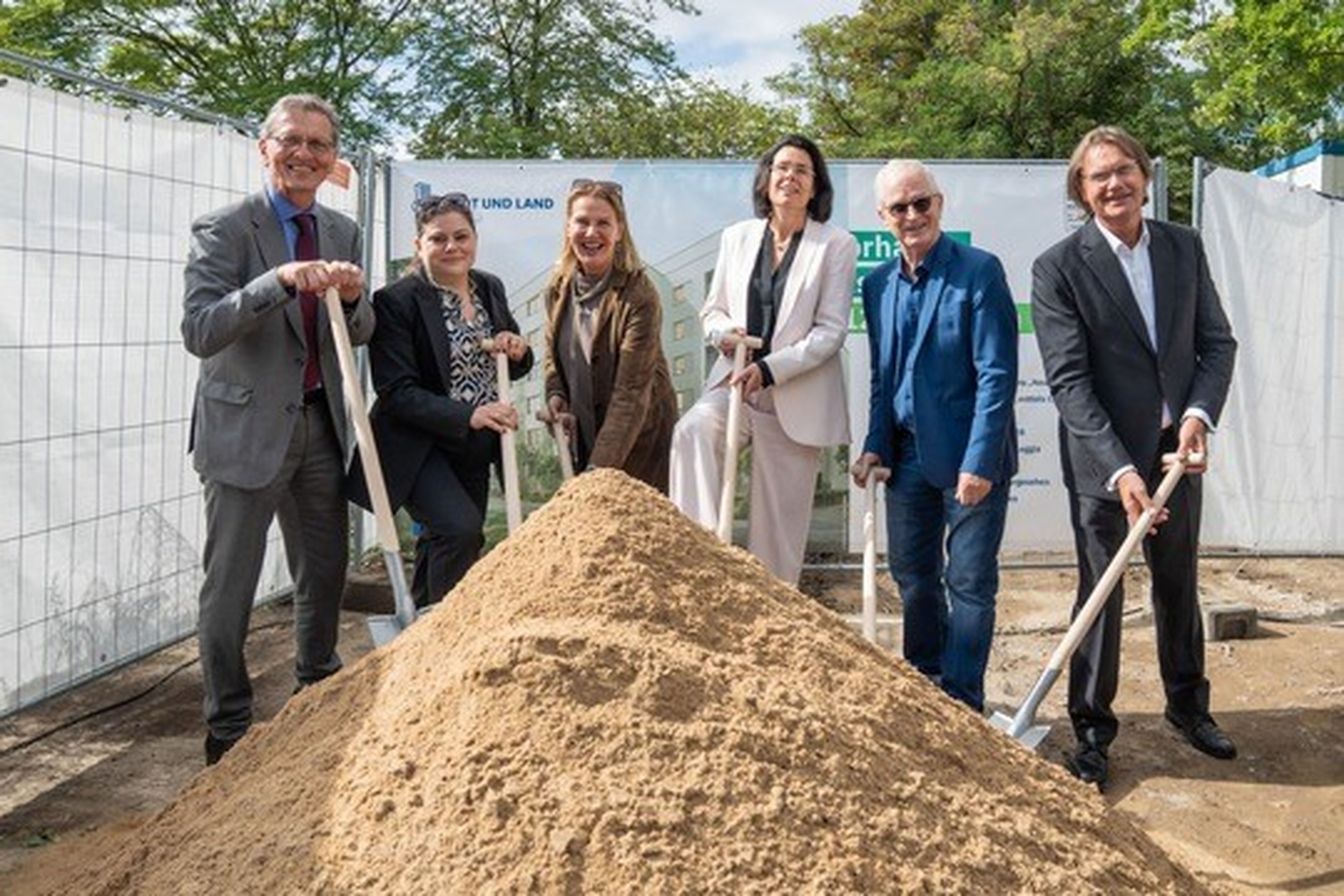 Six people in formal attire holding shovels stand behind a pile of sand at a groundbreaking ceremony with a fenced area and trees in the background.