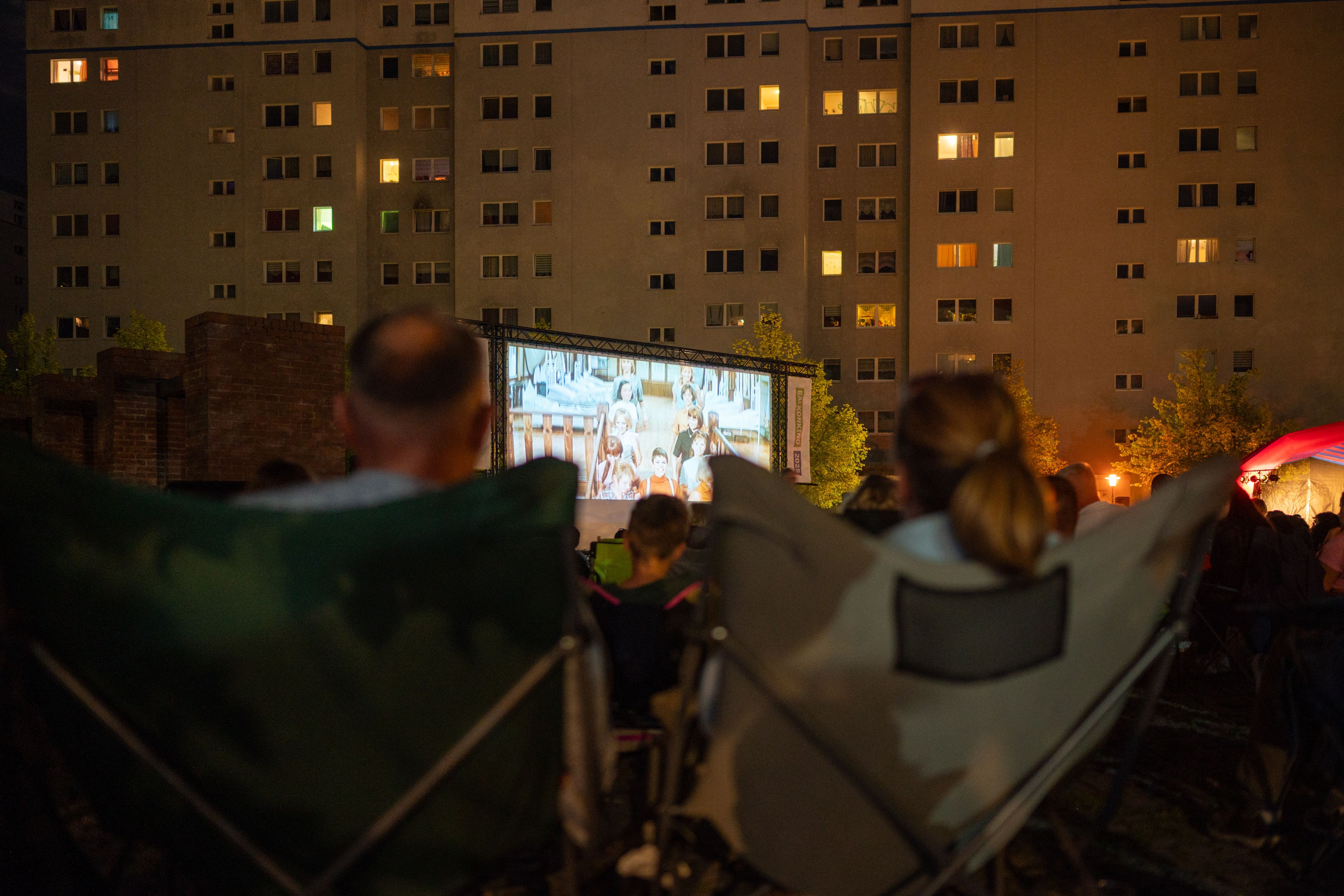 People sit in foldable chairs watching an outdoor movie screening in a city courtyard at night, with illuminated apartment windows nearby.