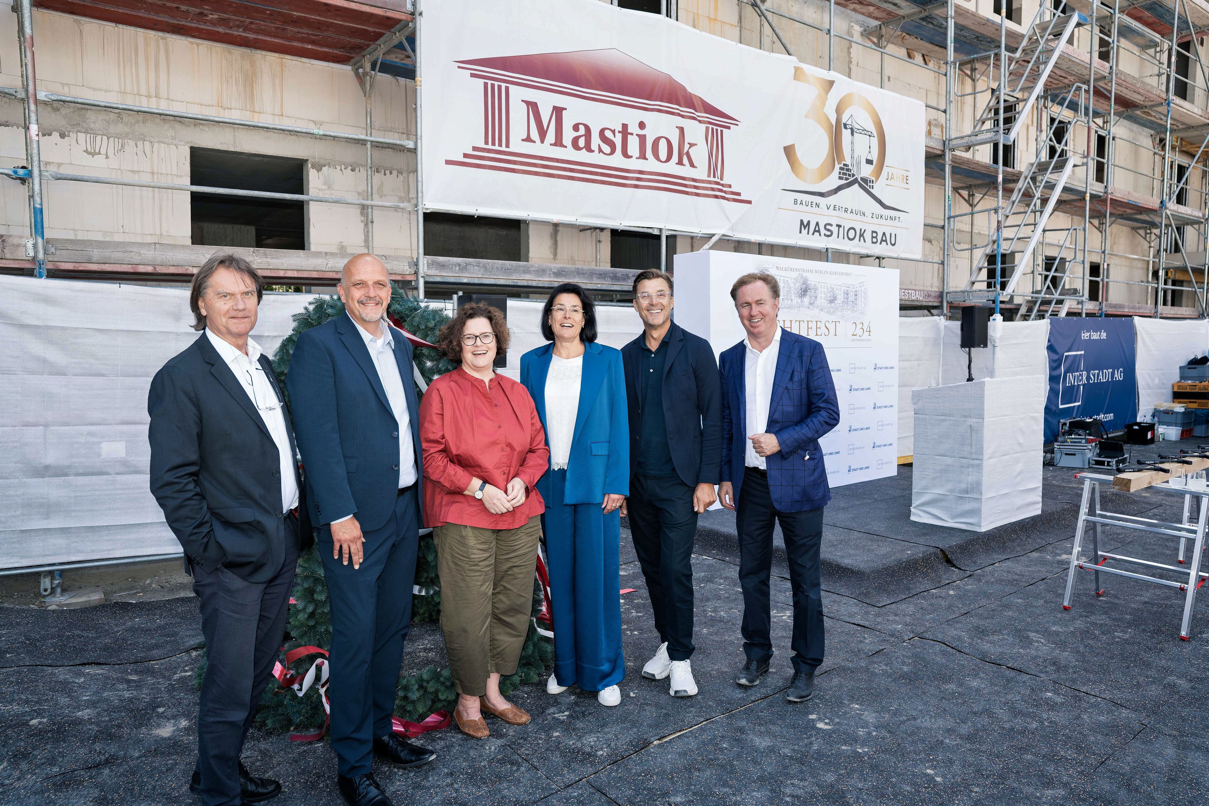 Five people standing together in front of a construction site with a "Mastiok" banner, celebrating a company's 30th anniversary.