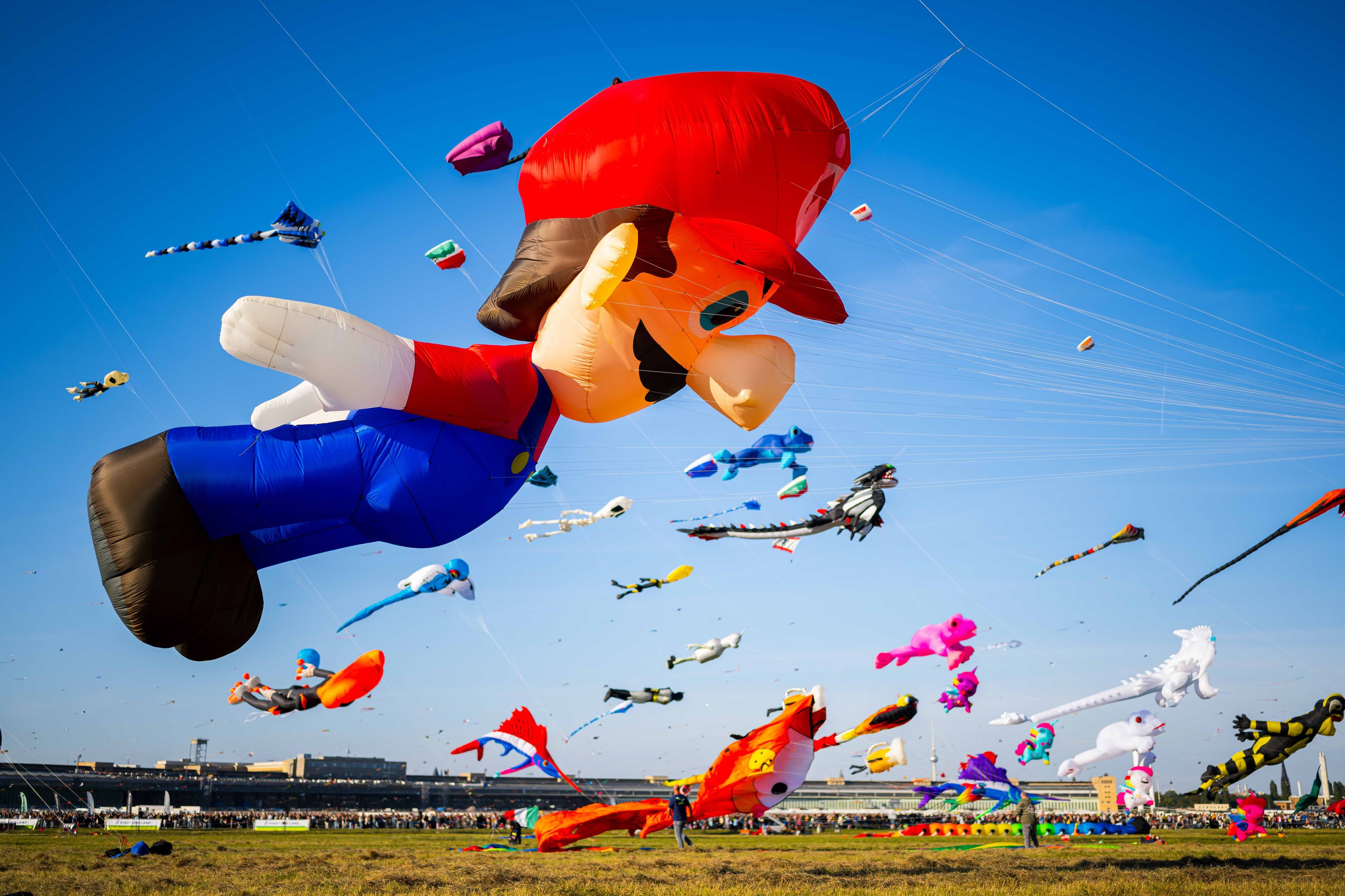 Large colorful kites shaped like cartoon characters and animals float in a clear blue sky during a kite festival with spectators below.