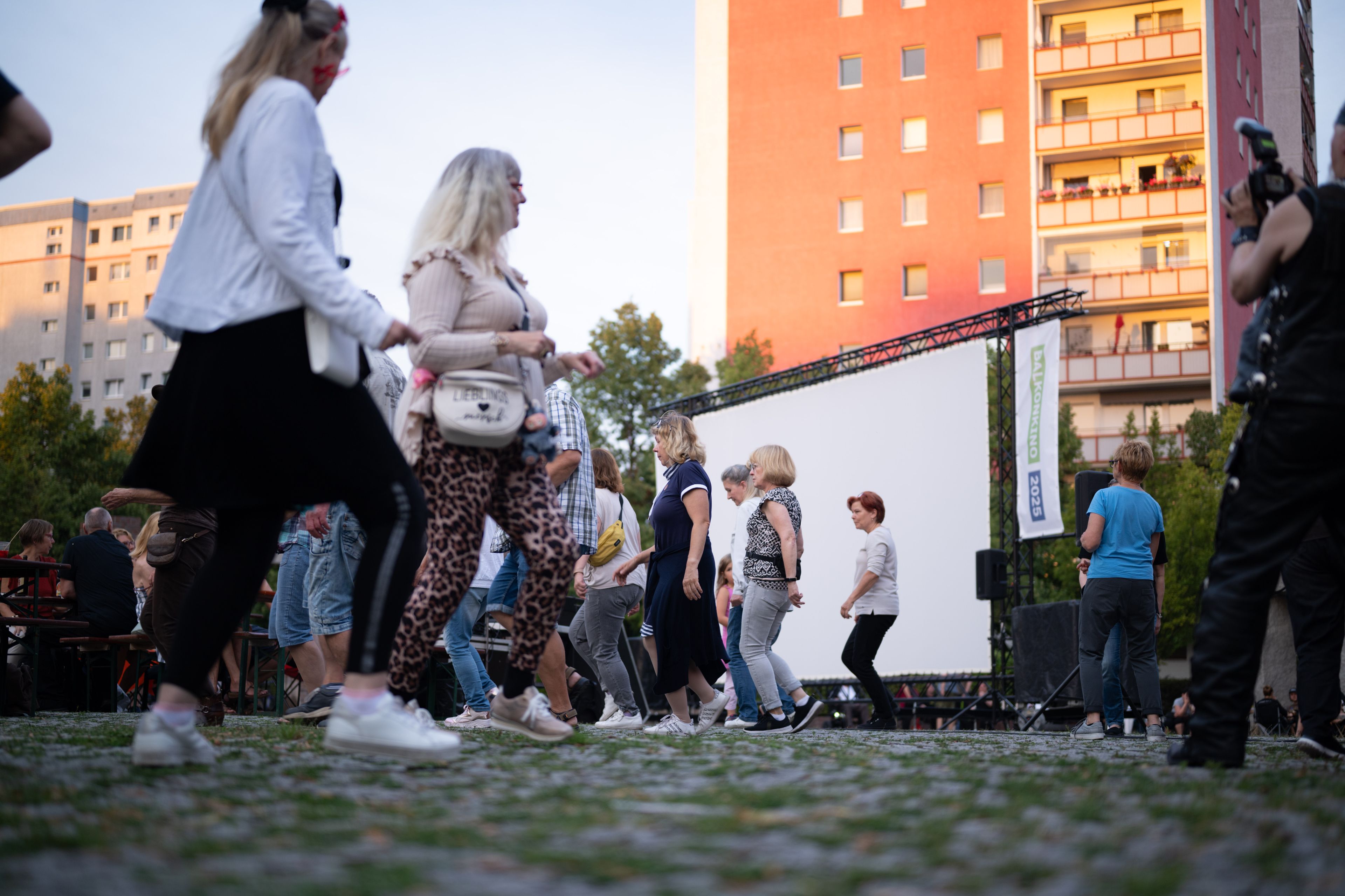 People dancing outdoors near a large screen, surrounded by tall buildings and trees, under a clear sky.