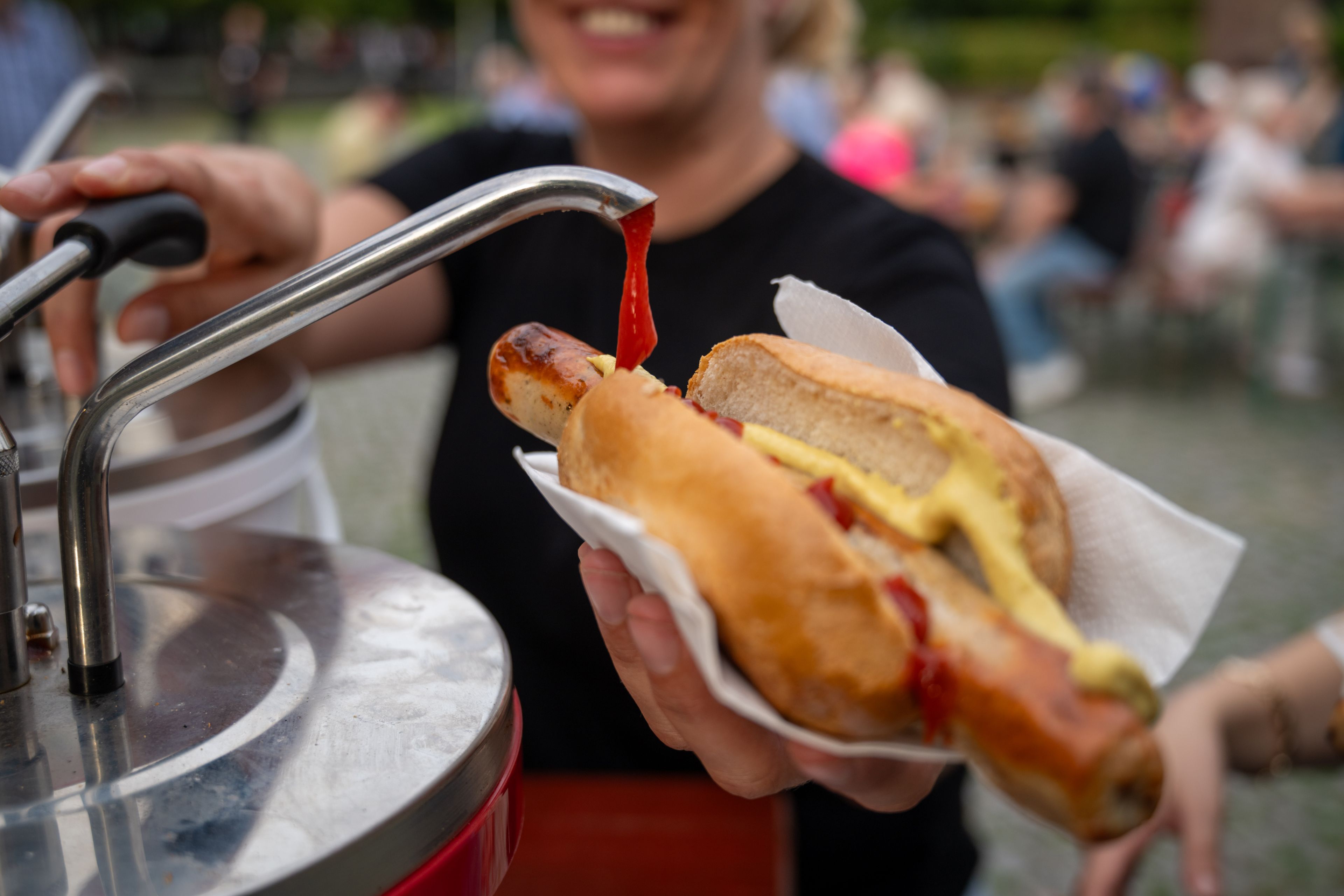 Person holding a hot dog with ketchup and mustard being dispensed onto it, smiling in the background.