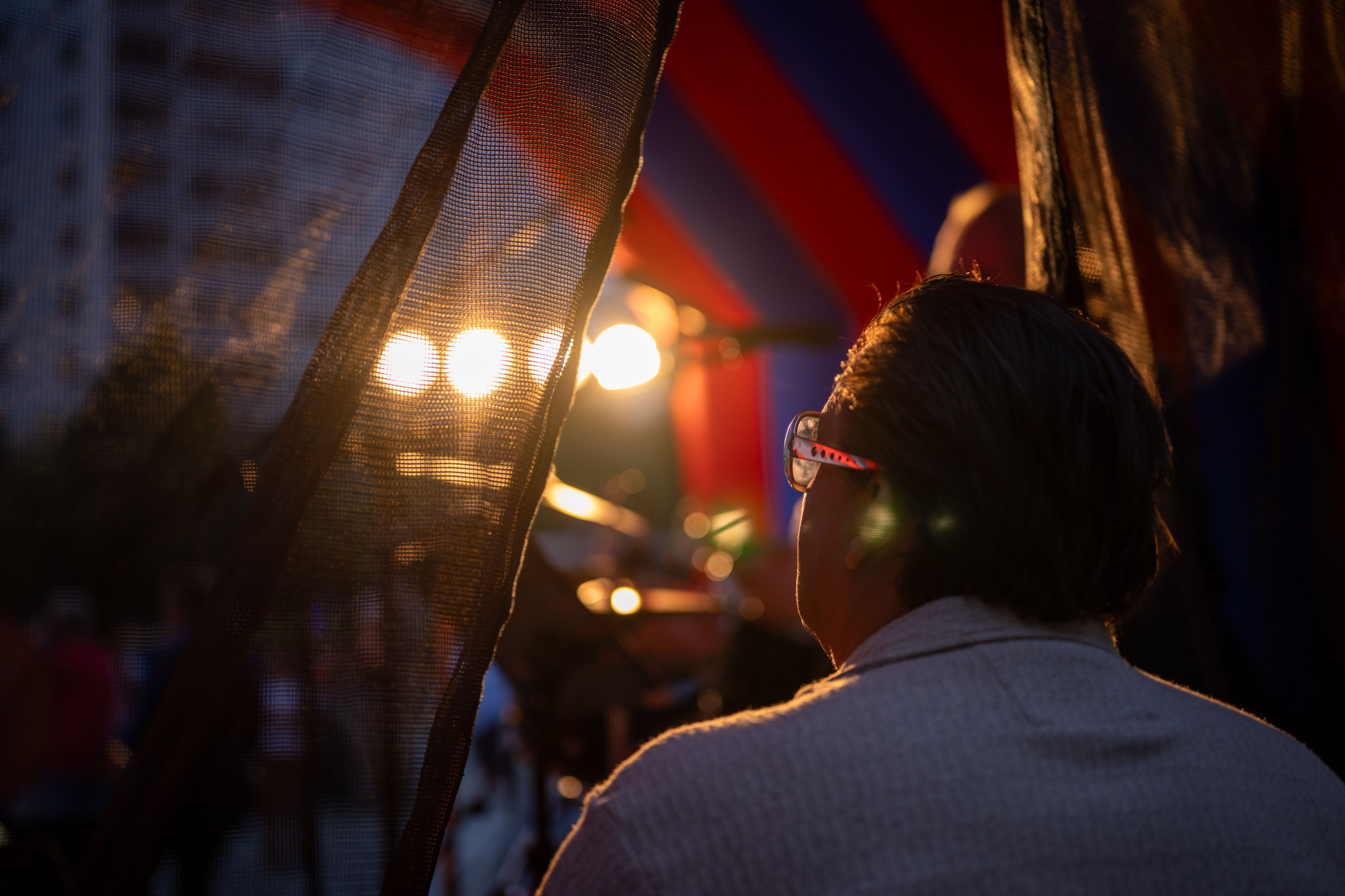 Person wearing glasses looks at stage lights through a curtain, with a colorful backdrop and an outdoor setting.