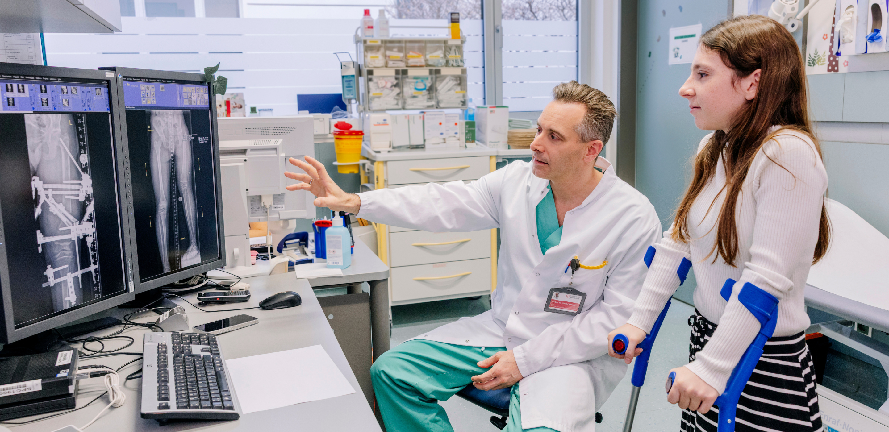 Ein Arzt zeigt einem jungen Mädchen mit Krücken etwas auf einem Bildschirm.