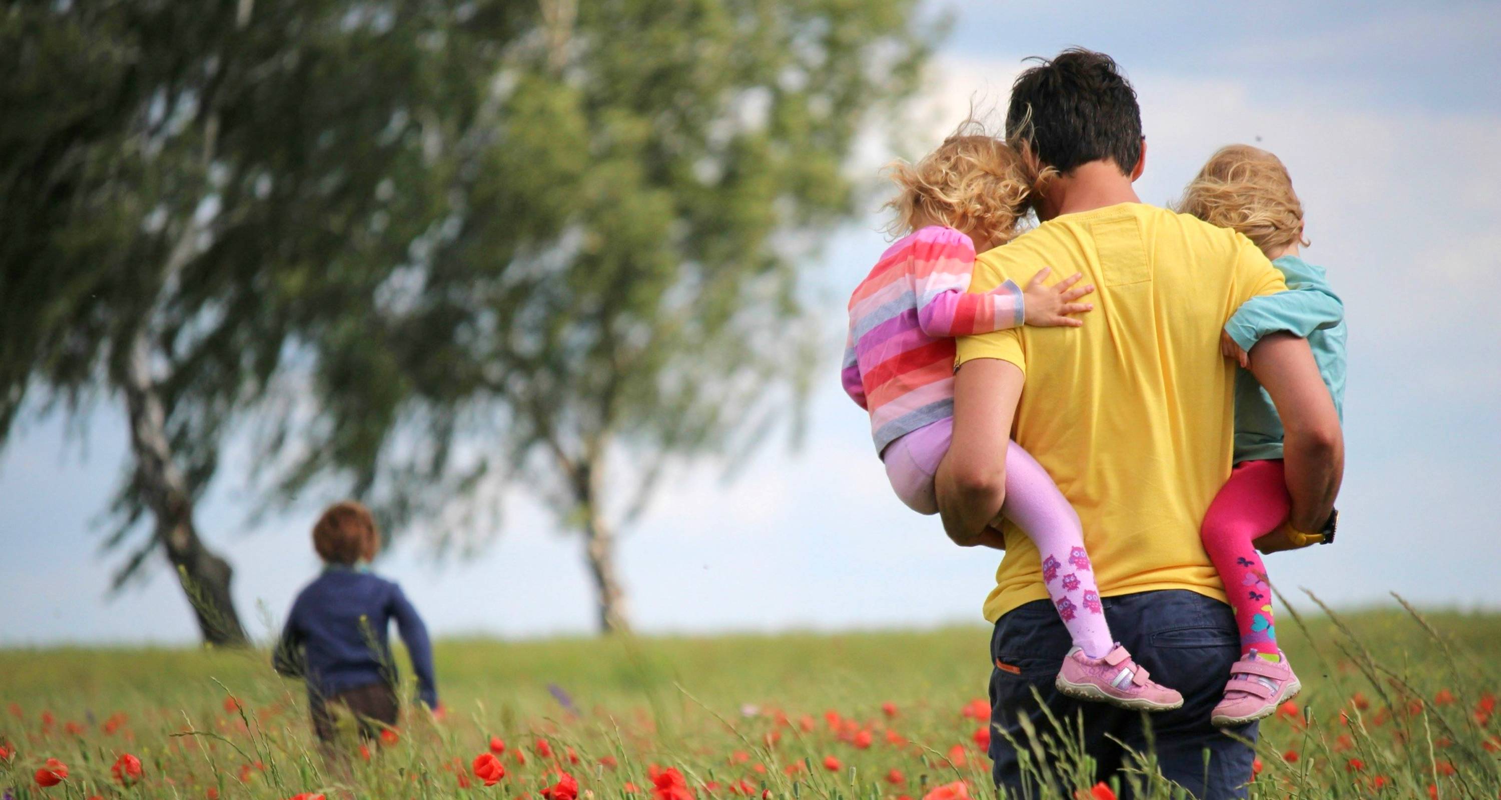 Ein Papa spaziert über eine Wiese und trägt dabei zwei seiner Kinder. Ein weiteres Kind läuft voraus.