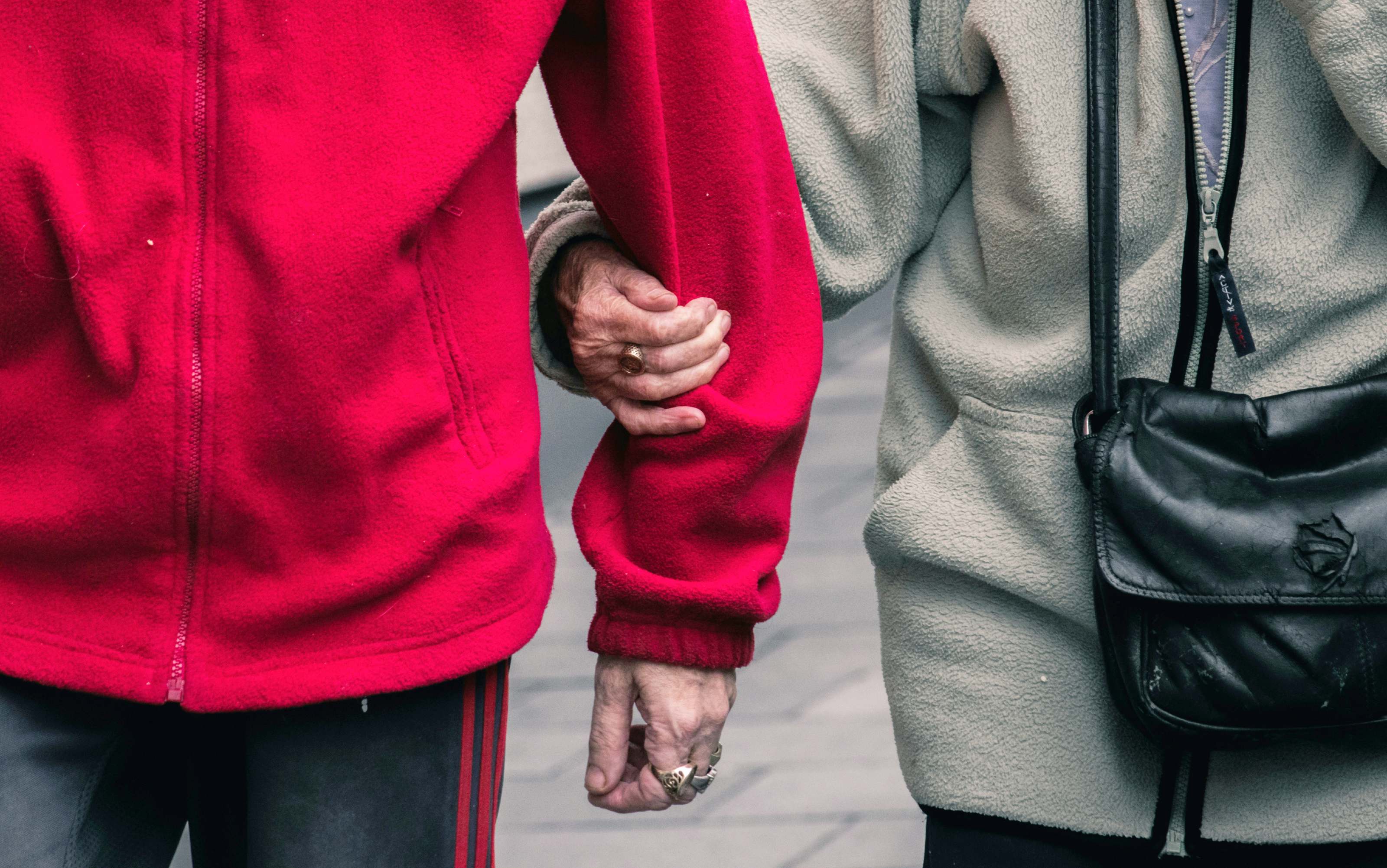 Eine Detailaufnahme von zwei älteren Personen. Eine von ihnen trägt einen roten Fleecepullover, die andere einen grauen. Sie halten sich am Arm.