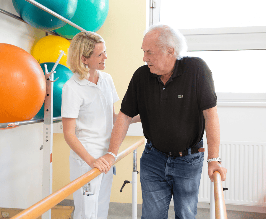 Älterer Mann stützt sich auf Gehhilfe, daneben Pflegerin